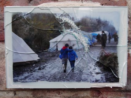Une photographie est recouverte d’un sous-verre brisé. Sur la photographie se trouvent deux enfants, de dos, qui marchent sur un chemin boueux. Le long du chemin, des tentes sont disposées.